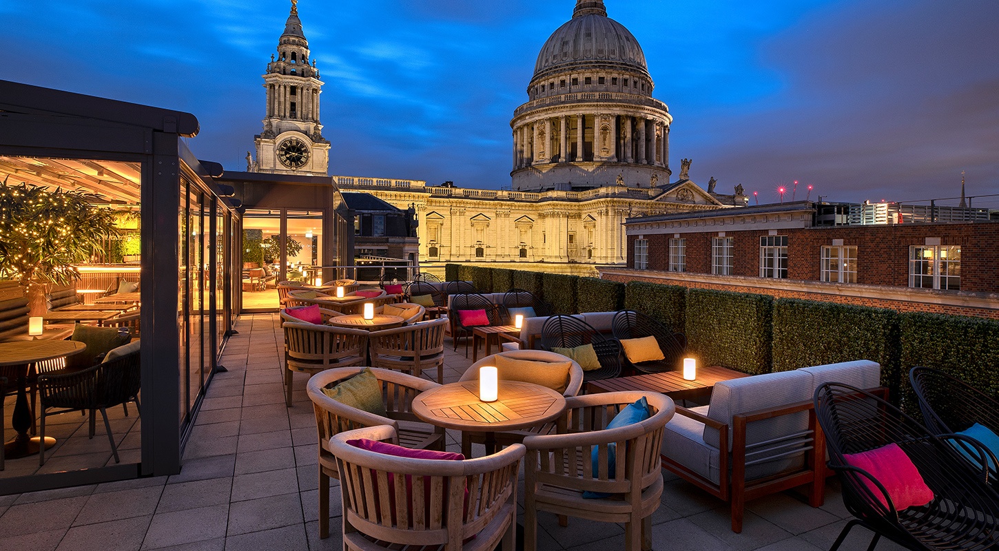 A photo of the Sabine rooftop at nighttime, with chairs and lit up tables on the roof seating area with St Paul's Cathedral visible in the background.