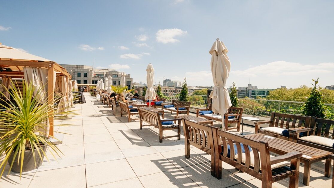 A photo of the Tower Suites rooftop in bright daylight, with benches and tables visible on the seating area, parasols and plants.